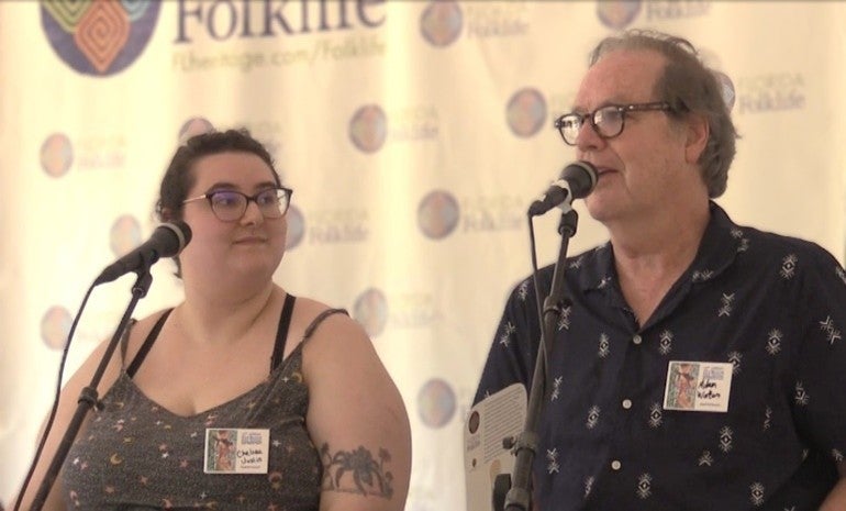 A man (Adam Watson) speaking into a microphone while a woman in front of a microphone (Chelsea Joslin) listens to him. They are presenting on a stage. 