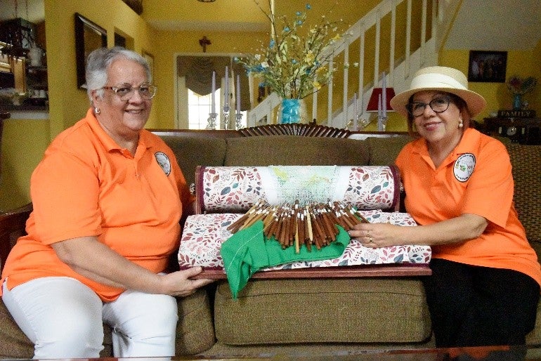 Two women in orange shirts sitting on either side of a mundillo box. 
