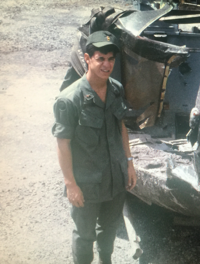 A man in military fatigues standing in front of a damaged vehicle