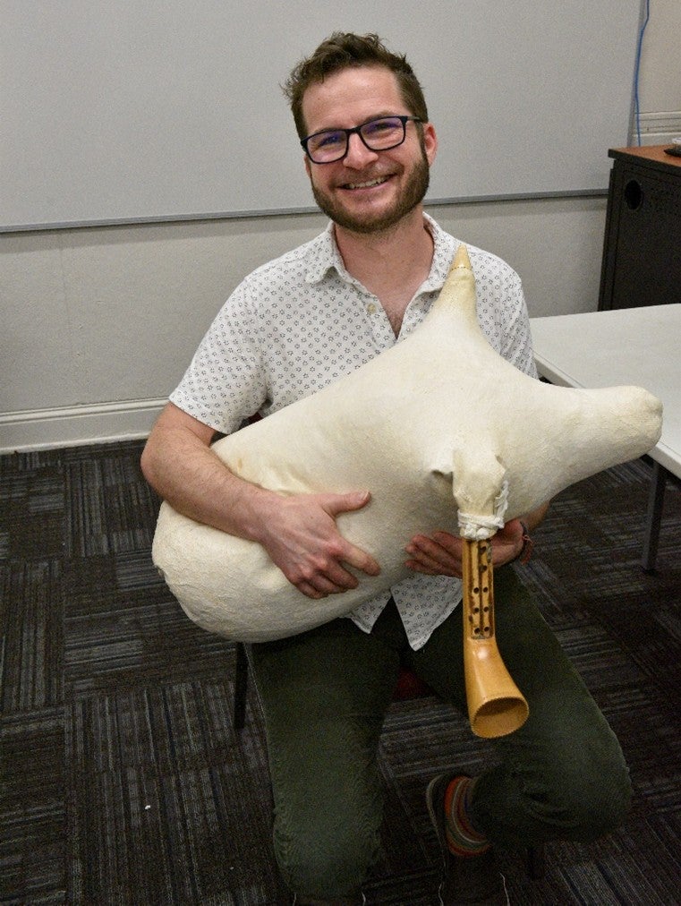 A bespectacled man holding a tsabouna (a cream-colored bag with a small wooden horn attached to it). 