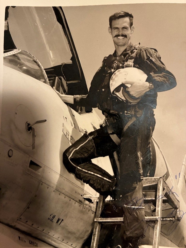A man wearing a flight suit stands next to the cockpit of a military plane
