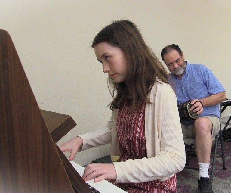 A girl in the foreground plays piano while a man behind her plays a concertina. 