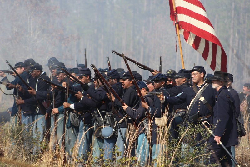 a line of african-american reenactors in union dress form a firing line with an american flag