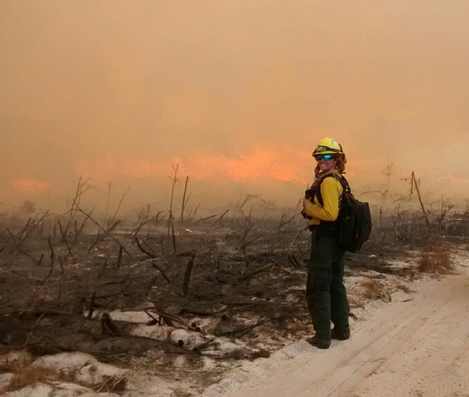A lady with fire gear on stands infront of land that has just been burned. 