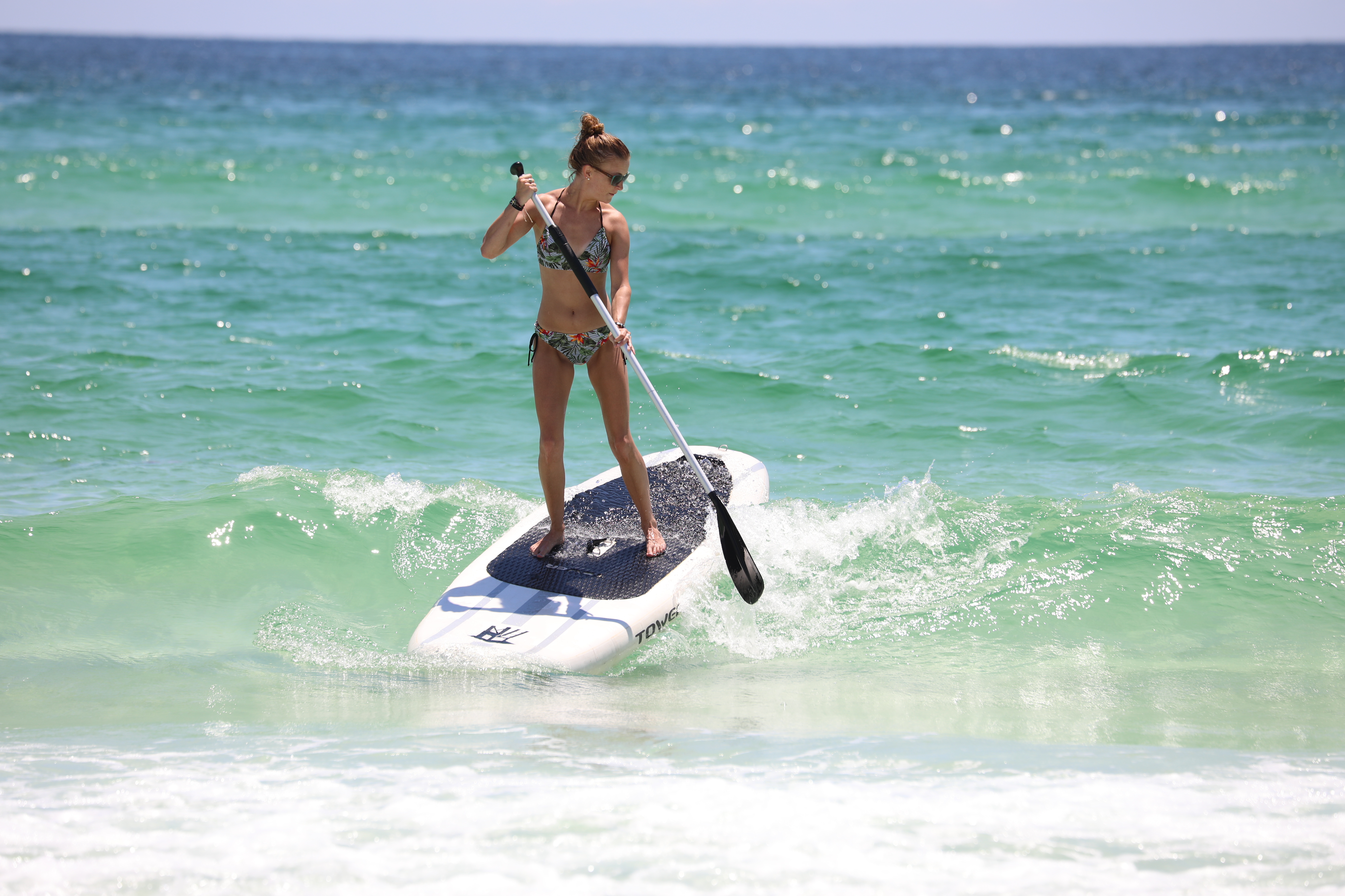 Woman standing on paddle board rides a wave in the emerald green surf. 
