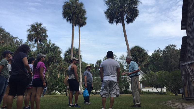 a park ranger in period clothing talks to a large tour group outside underneath palm trees
