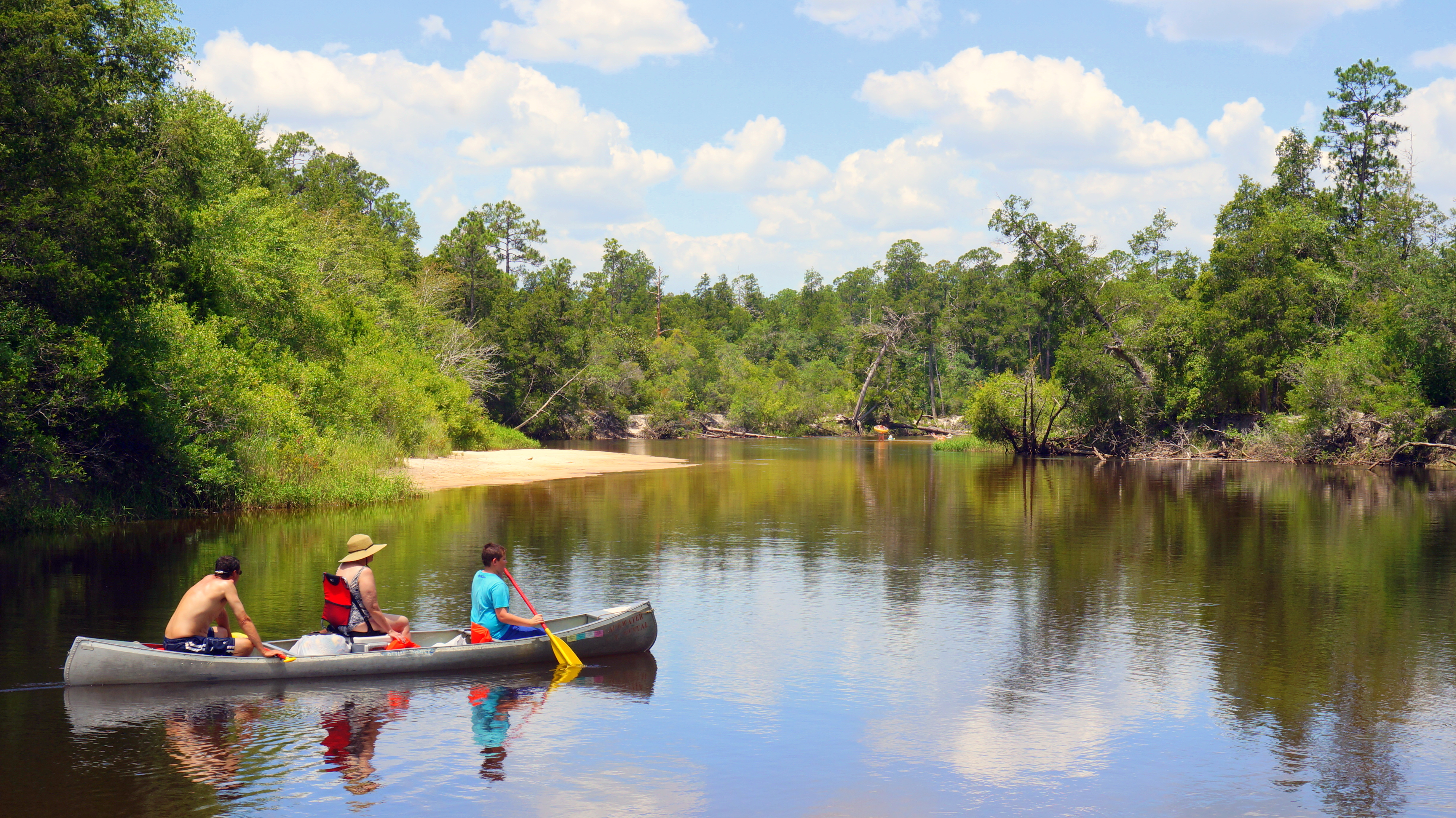 Three people in a canoe glide through tannin waters reflecting the clouds and sky. 
