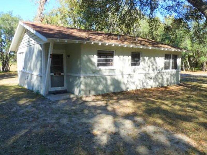 a white block cabin with a brown roof sits under the shade of trees