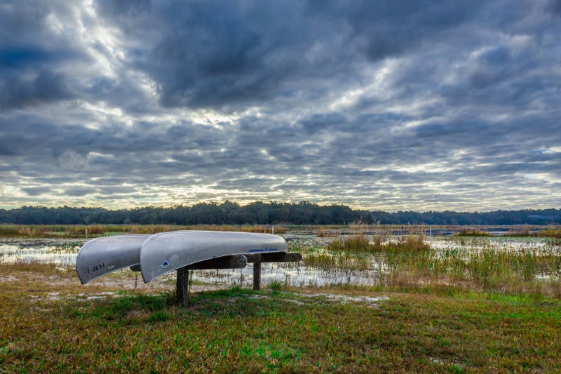 two canoes stand on a wooden platform in from of a lake, and above a sky streaked with clouds.