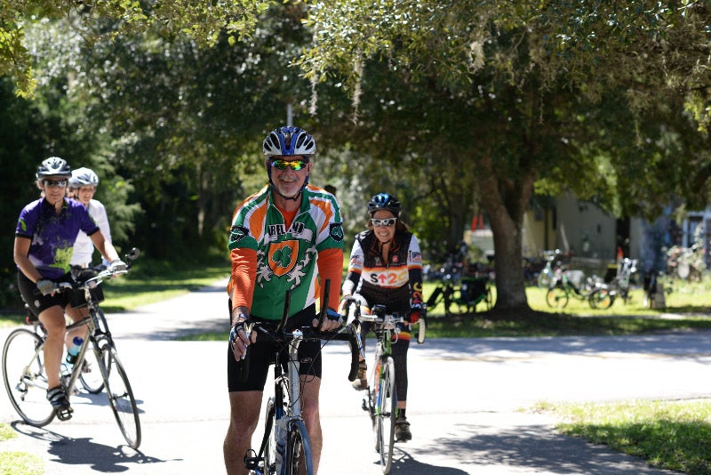 four cyclists beam at the camera from a sunny paved path.