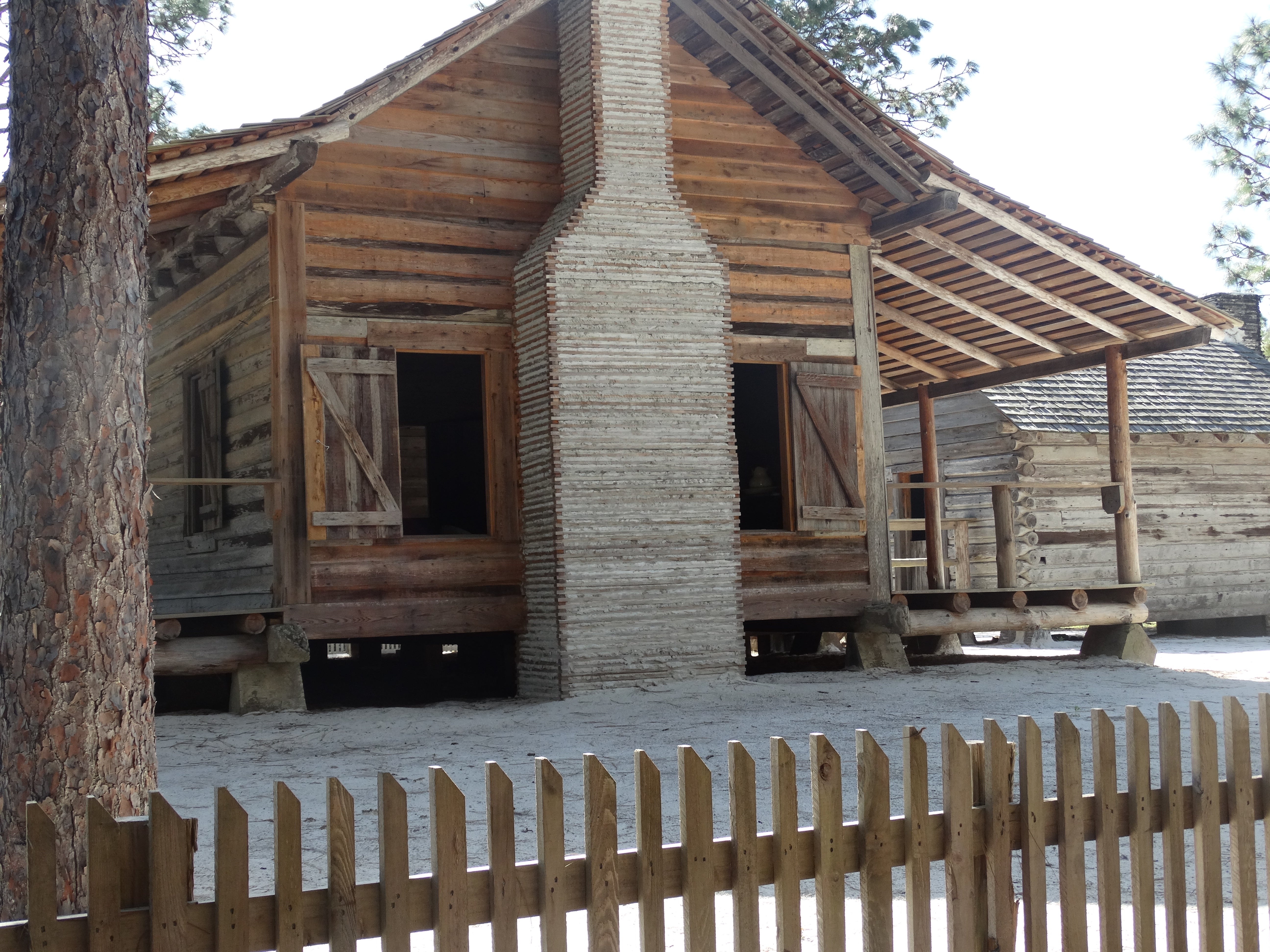 a log cabin with a large chimney sits behind a wood picket fence.