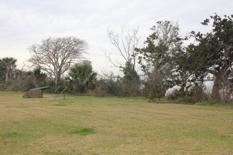 a grassy field with a cannon, signs, and trees overlooking a river.