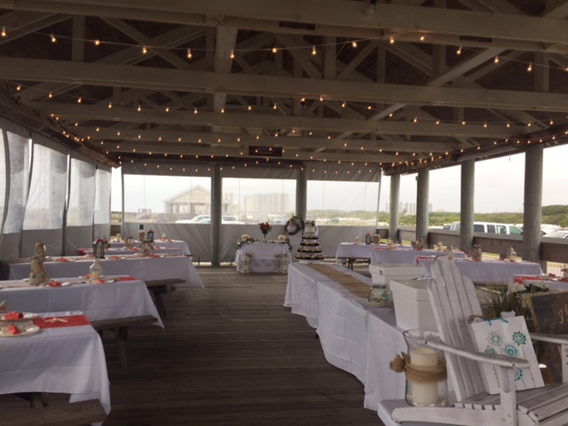 Picnic tables are covered with white table cloths and decor.  Soft bistro lights are draped throughout the pavilion.