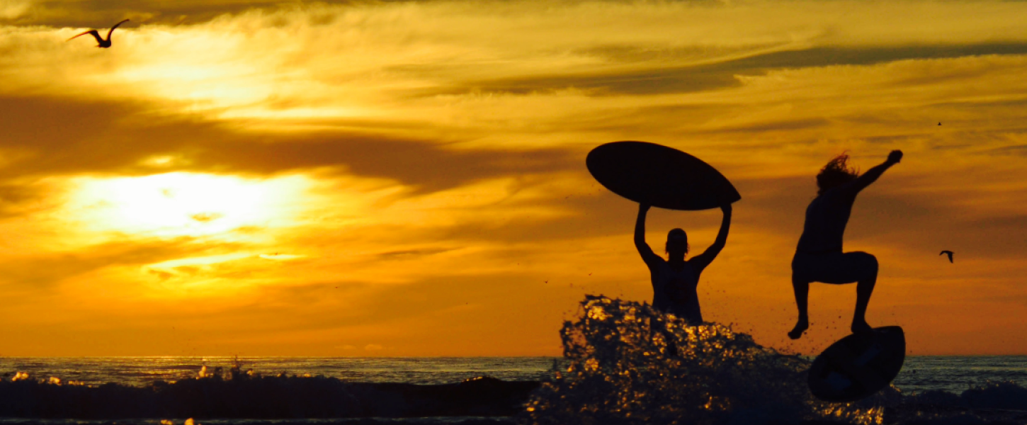 A silhouette of two people playing with skim boards in the surf with a golden yellow sky behind them.