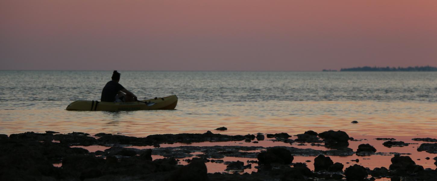 A person sitting in a kayak in the ocean, looking out at the sunrise. Small silhouetted rocks are in the foreground. 