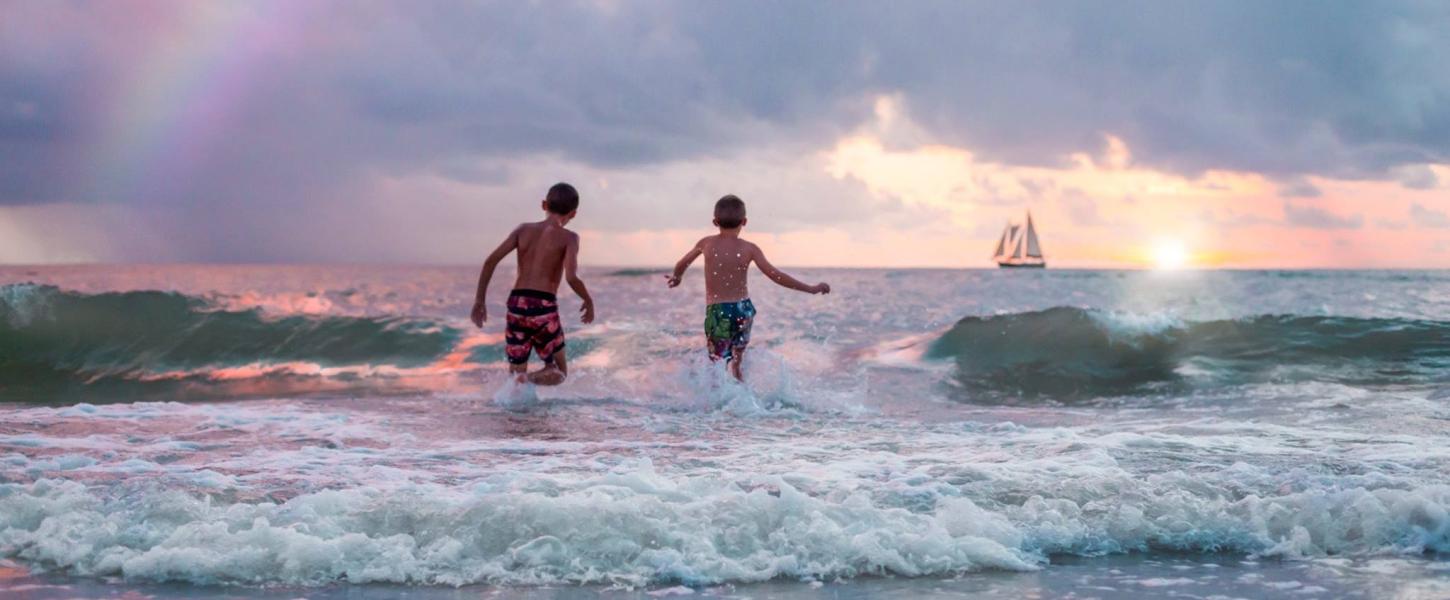 Two teenage boys running into the waves in the late afternoon, framed by a distant sailboat and part of a rainbow.