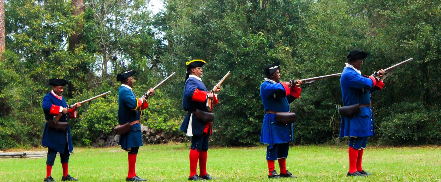 A line of 5 reenactors wearing blue and red historic costume prepare to fire muskets. 