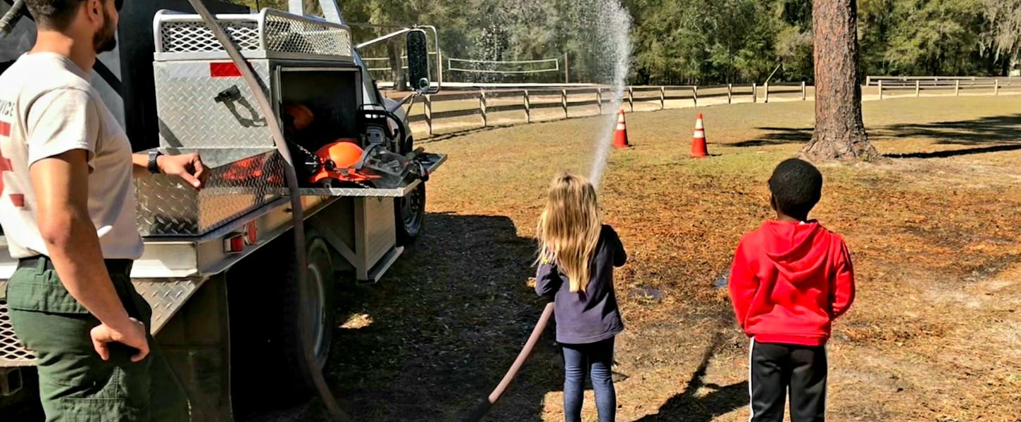 A young girl spraying water from a fire hose while a young boy and a Ranger watch.