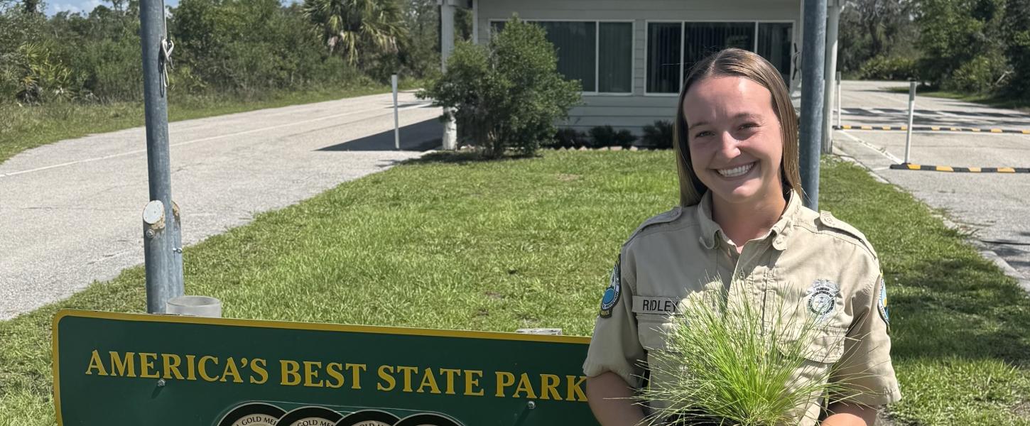 A smiling woman in park ranger uniform, holding a pot of long grass. 