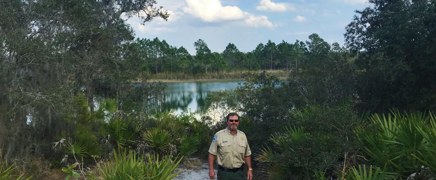 Park Manager Andy Noland standing at Catfish Creek surrounded by saw palmetto, oaks, and a lake behind him