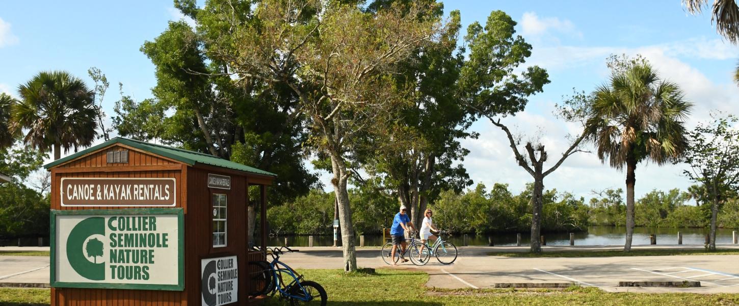 A man and a woman riding bicycles. River water in background. Rental building to the left.