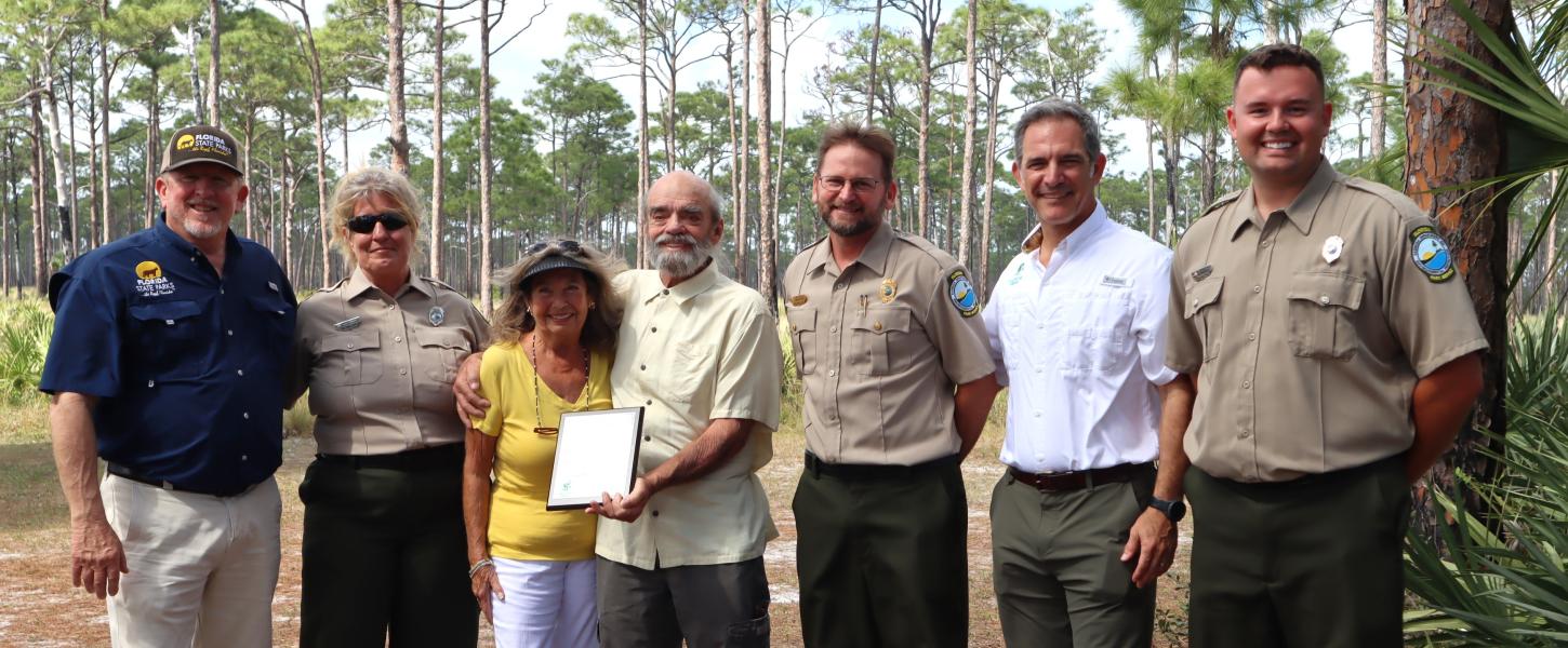 A group of people standing shoulder to shoulder. A man and woman in the middle are holding a plaque. 