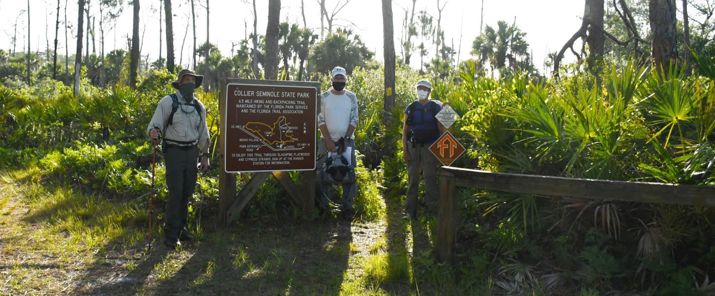 Three hikers standing by a hiking trail sign with green trees and grass.