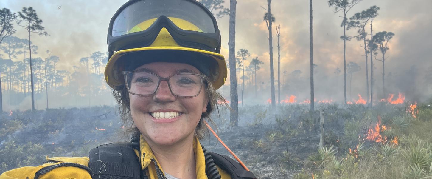 A smiling woman in fire PPE, standing in front of a prescribed fire. 