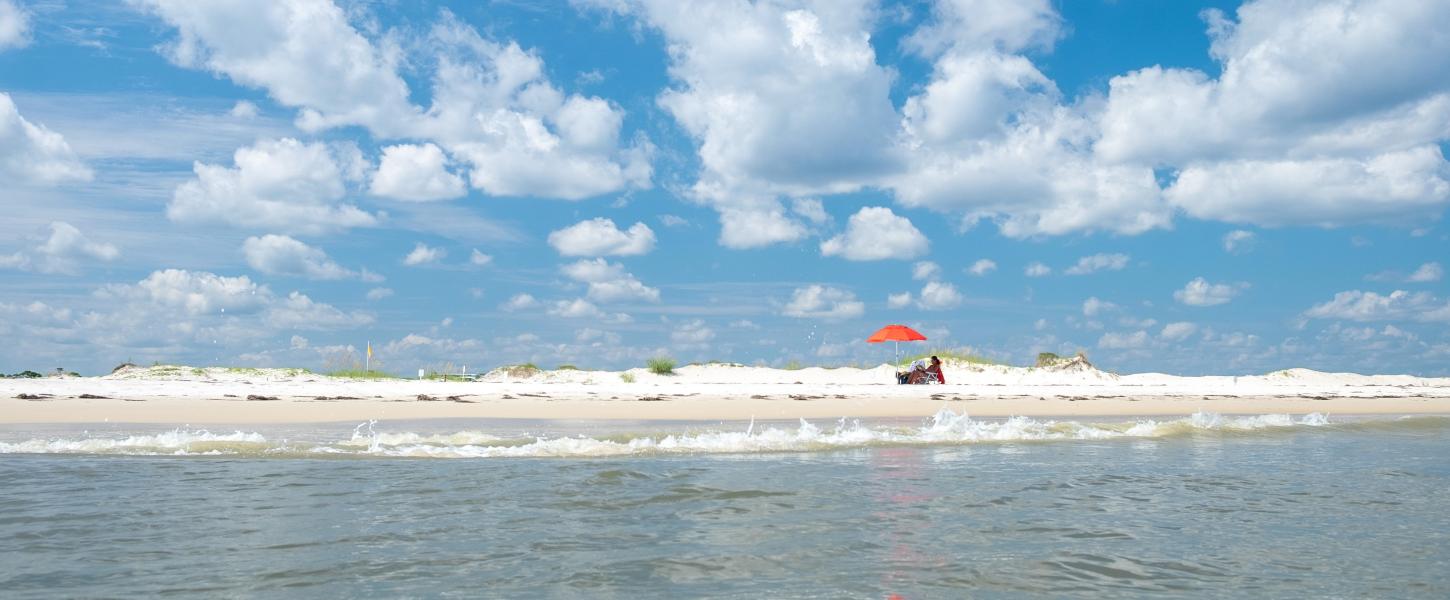 A person sits in a chair with an orange umbrella on the beach at St. George Island State Park.