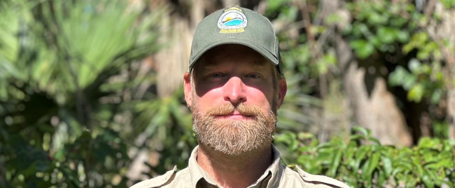 A bearded park ranger standing in front of greenery. 