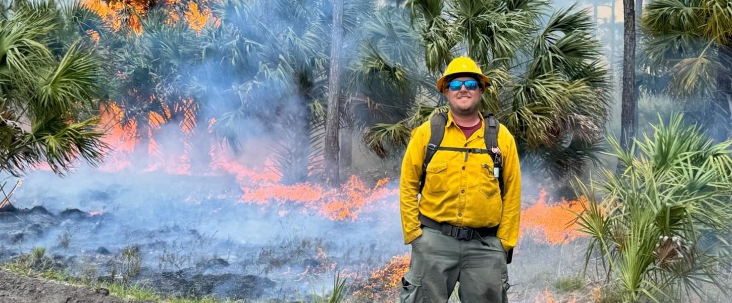 A smiling man wearing protective equipment standing in front of a large prescribed burn. 