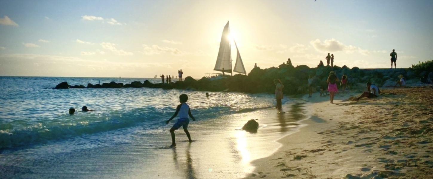 a beach cove with people playing while a sailboats passes in the background. 