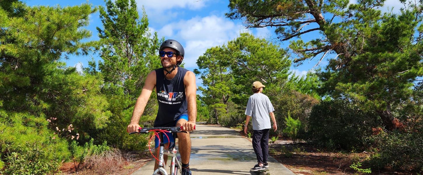 Biker and skateboard on paved path through trees. 
