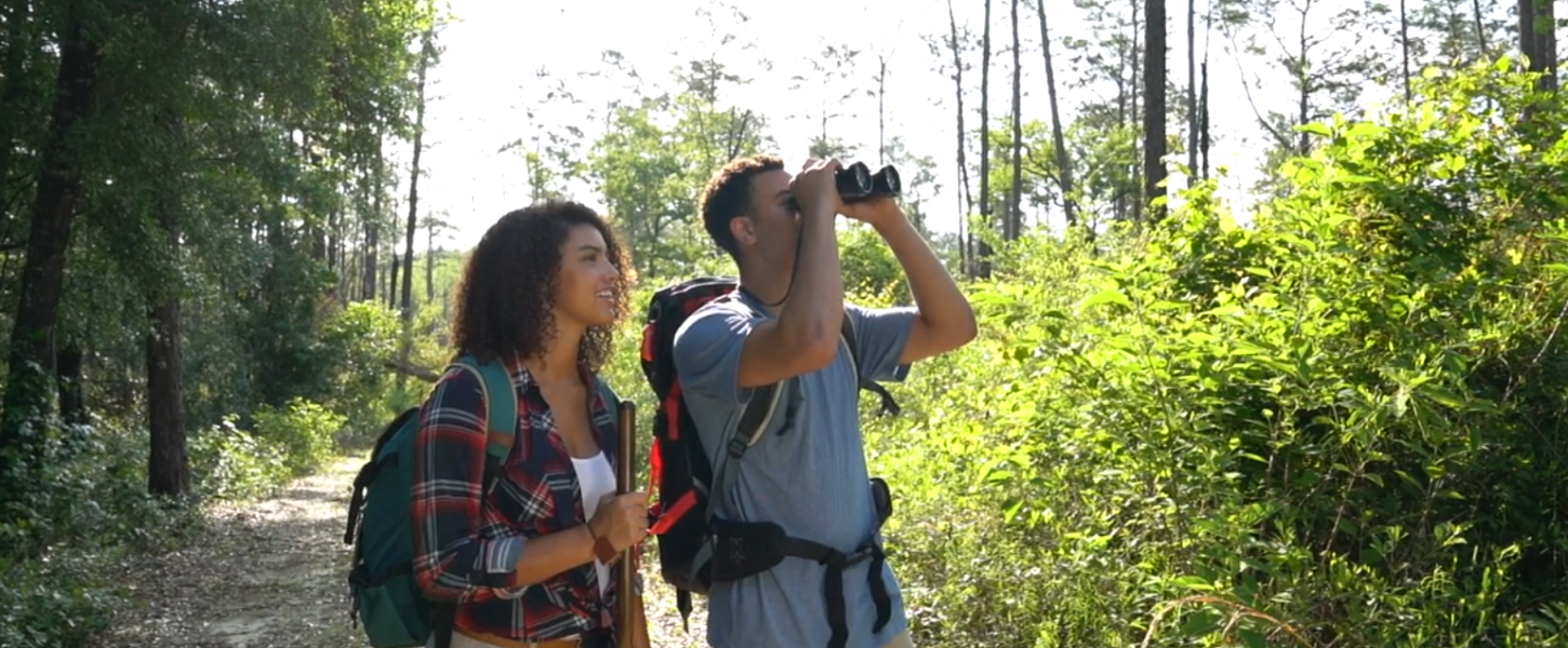 A couple in the woods, one looking through a pair of binoculars. 