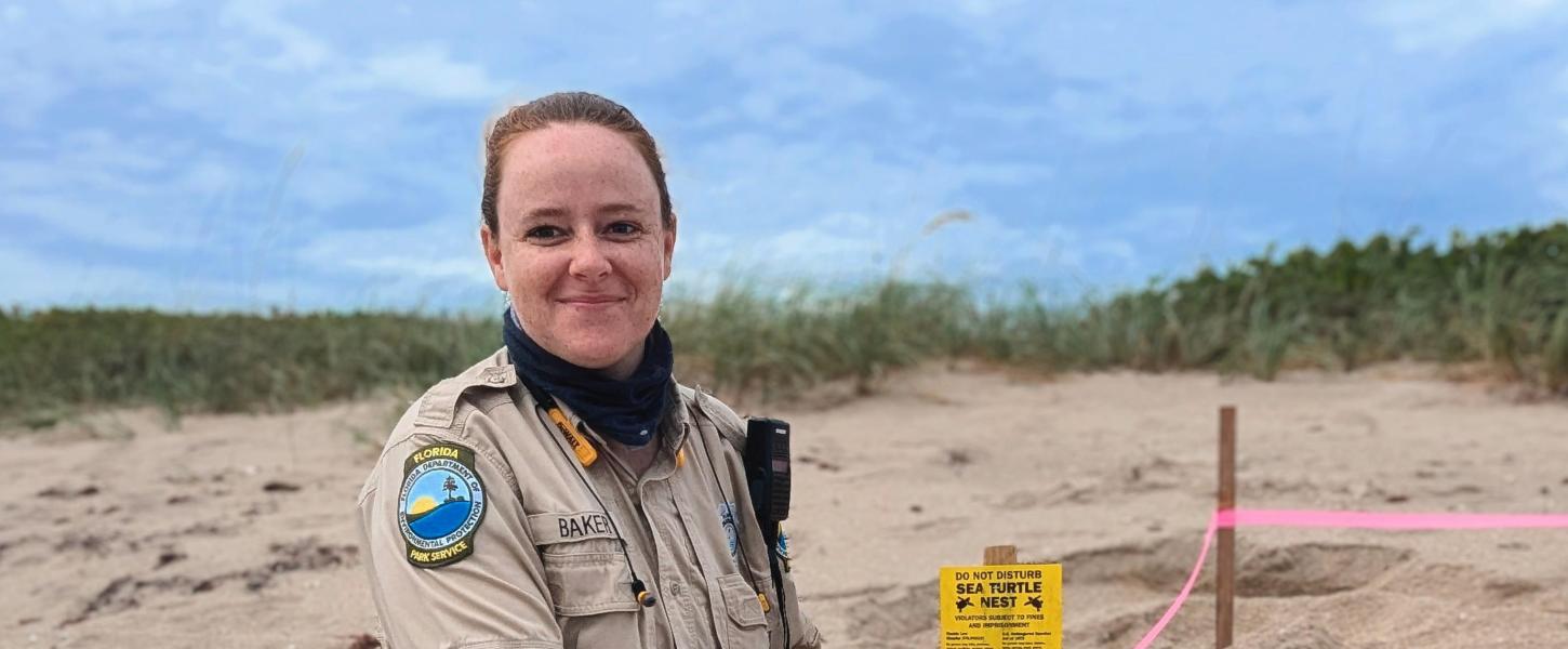 A smiling park ranger writing on a clipboard next to a sea turtle nest.