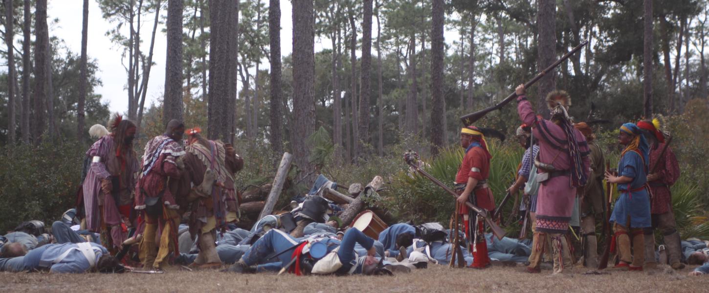 Seminole reenactors in colorful garb raise their rifles in celebration after their victory of the soldiers.