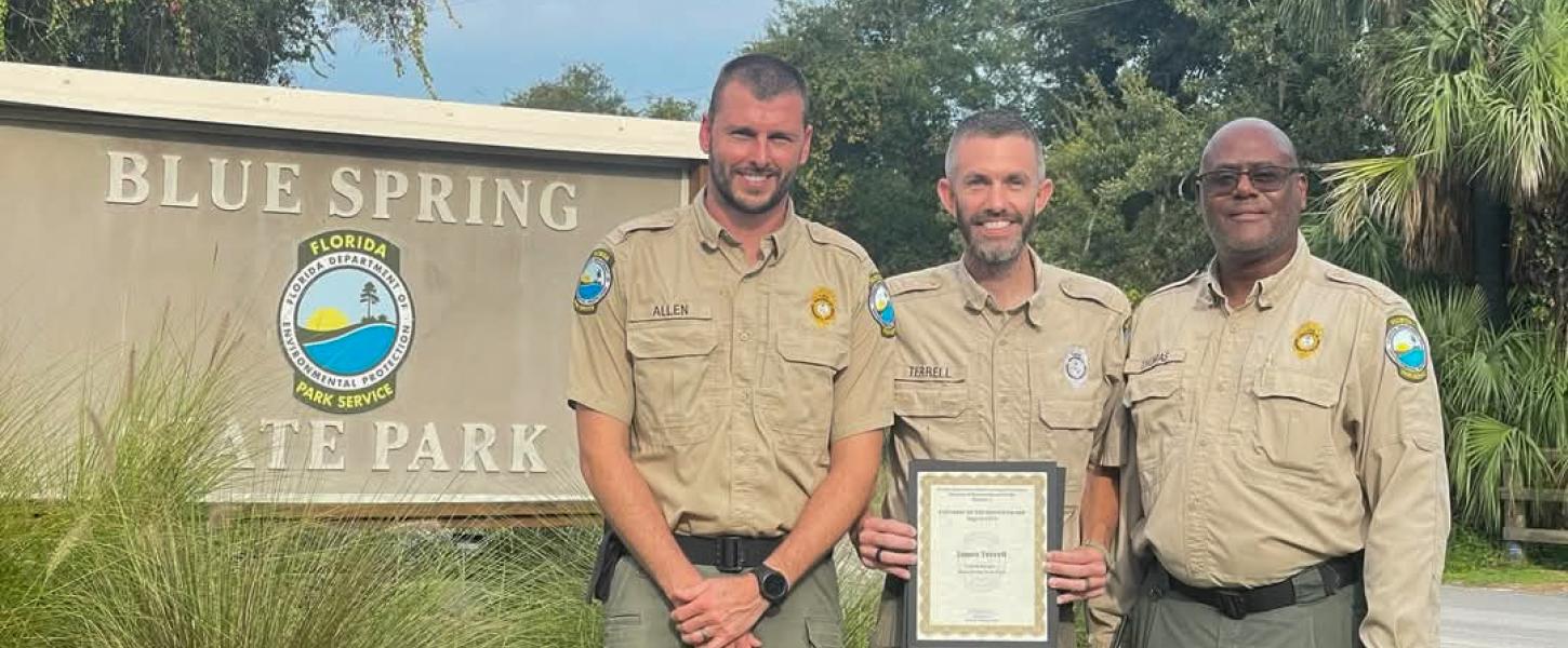 Three park rangers standing in front of a sign that reads "Blue Spring State Park". The park ranger in the middle is holding a framed certificate.  