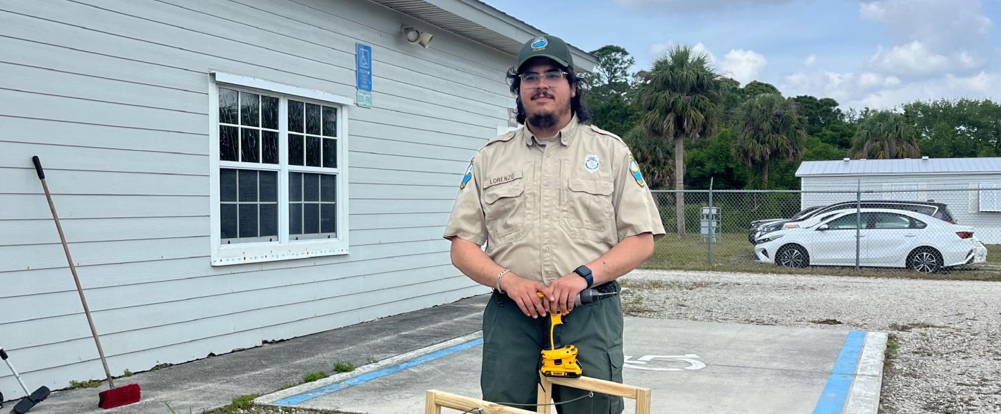 A park ranger holding a drill, standing over a wooden project. 