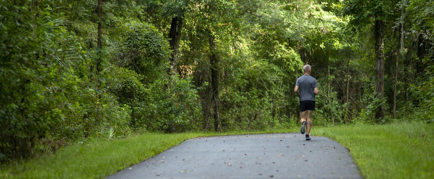 A man running on a paved trail through a lush green forest. 