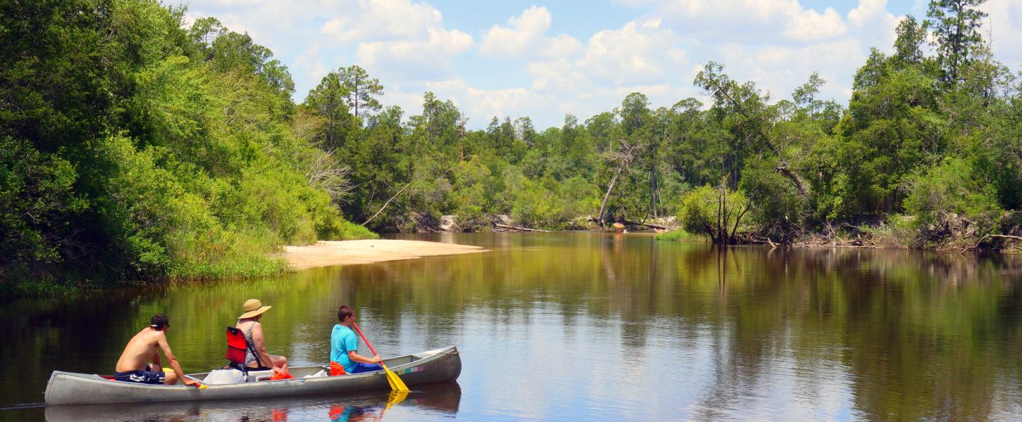 Three people in a canoe glide through tannin waters reflecting the clouds and sky. 