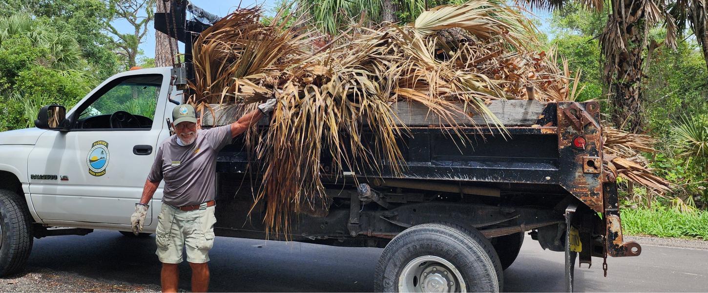 A man standing in front of a pickup with his hand on a pile of palm leaves that is taking up the bed of the pickup. 