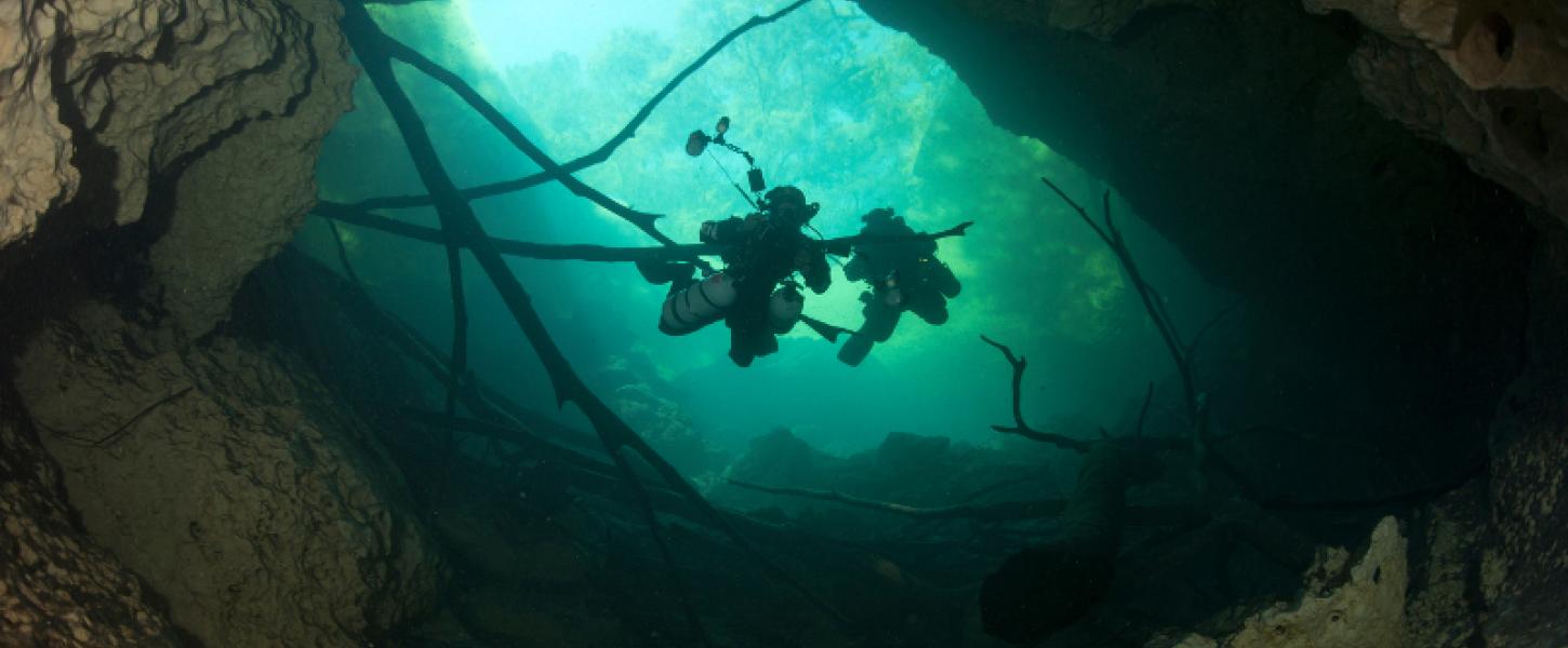 Two divers descend down into an underwater cave, backlit by a blue-green light