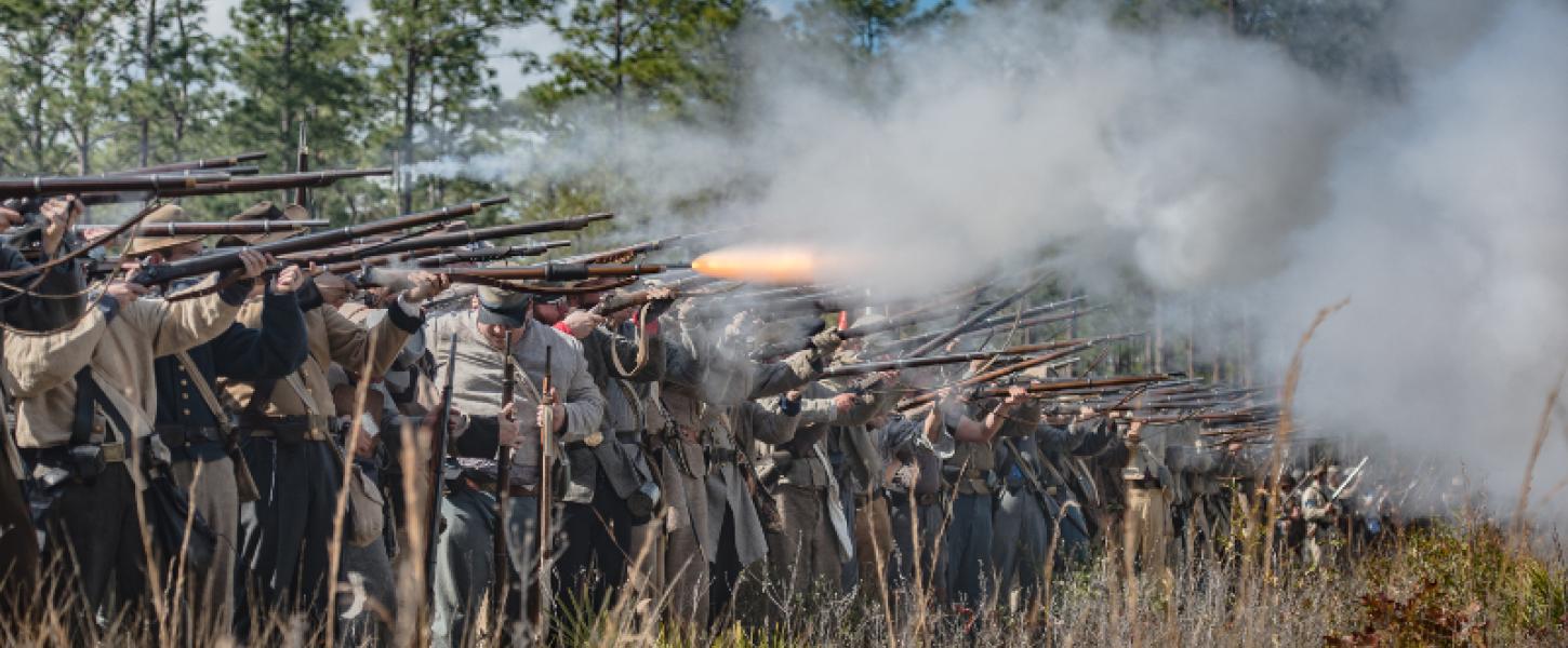 reenactors dressed as confederate soldiers stand in a long line firing guns