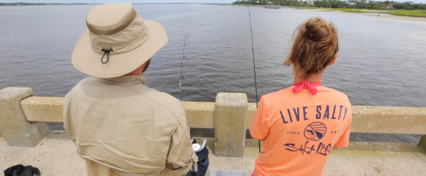 a man and woman with backs to the camera fish off of a concrete bridge