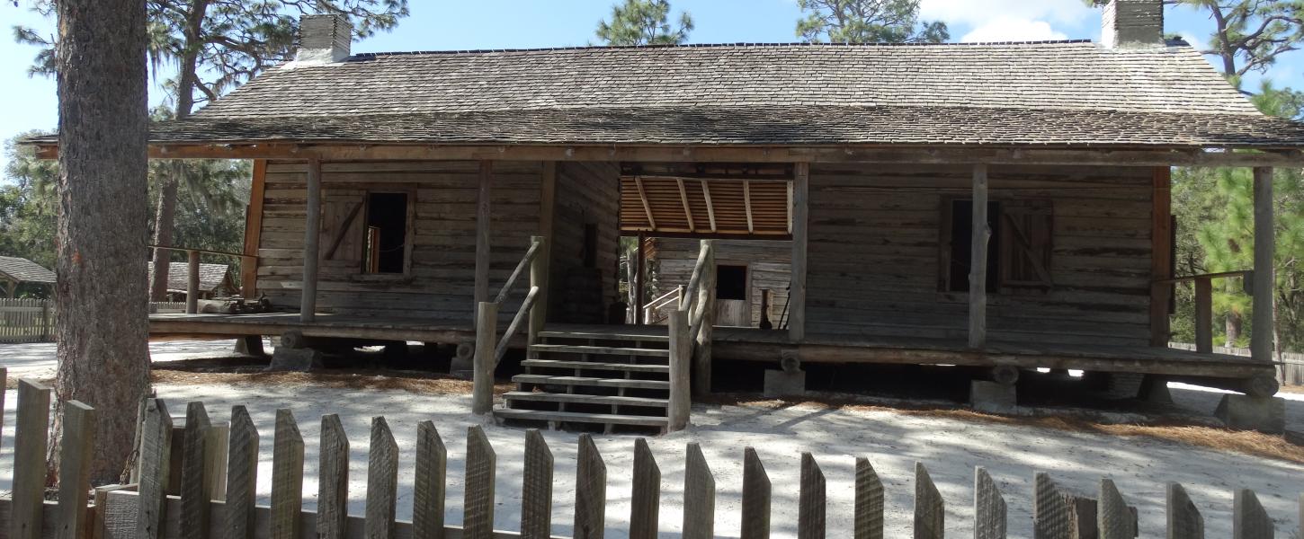 a log cabin with a wood picket fence sit under a blue sky surrounded by pine trees