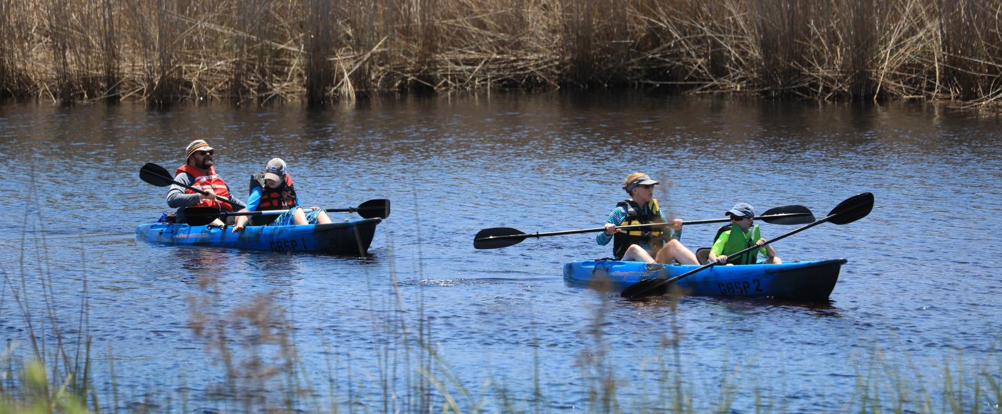 Two kayaks, each with an adult and a child, paddle on the coastal dune lake. 