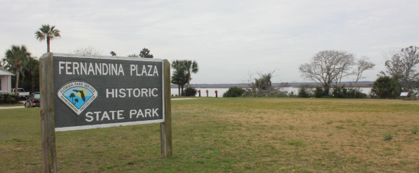 a sign reading fernandina plaza historic state park stands in a grassy field by a river