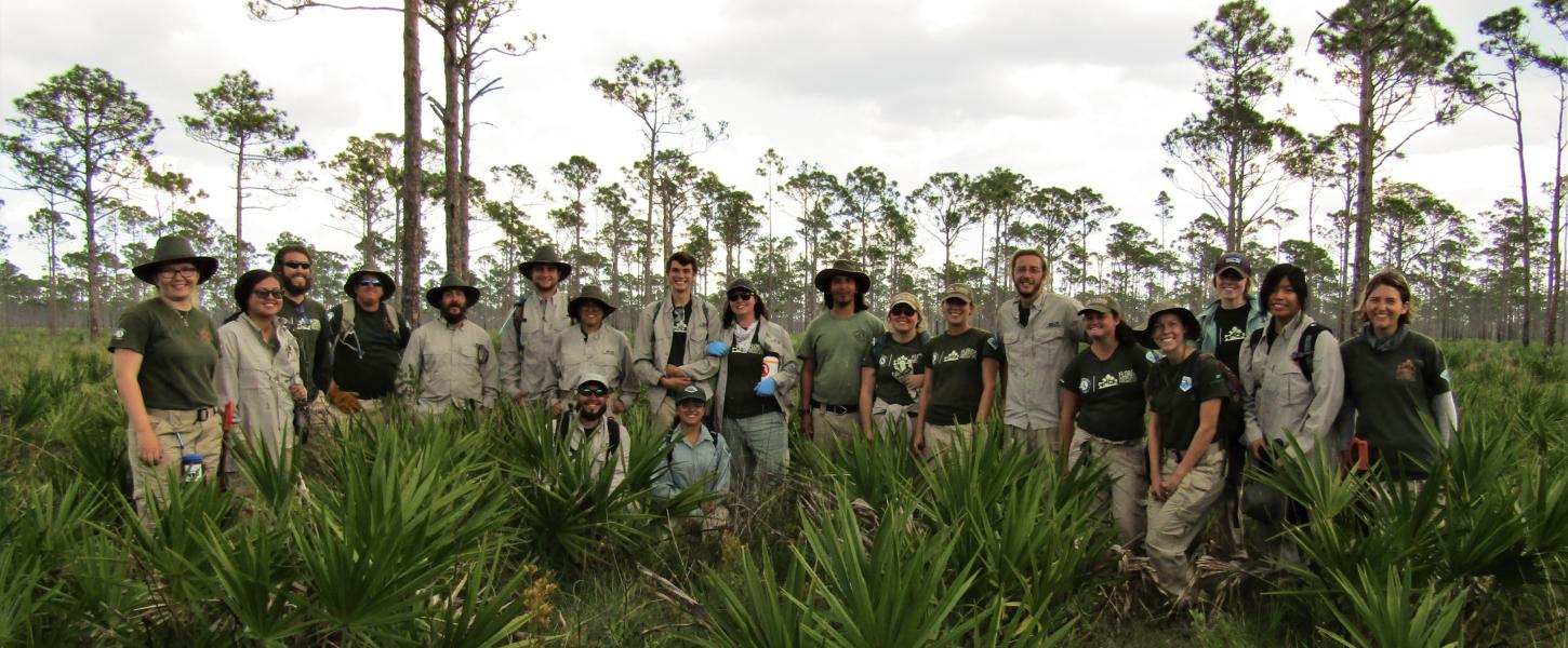 A group of FLCC members pose in a pine scrub landscape, wearing khaki and green uniforms and smiling. 