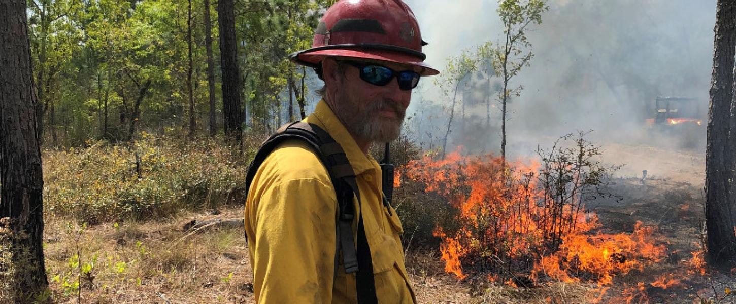 a man in protective gear stand sin front of a prescribed fire with a drip torch