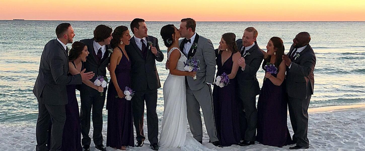 Wedding party surround bride and groom on white sandy beach with glowing orange sunset in the background.